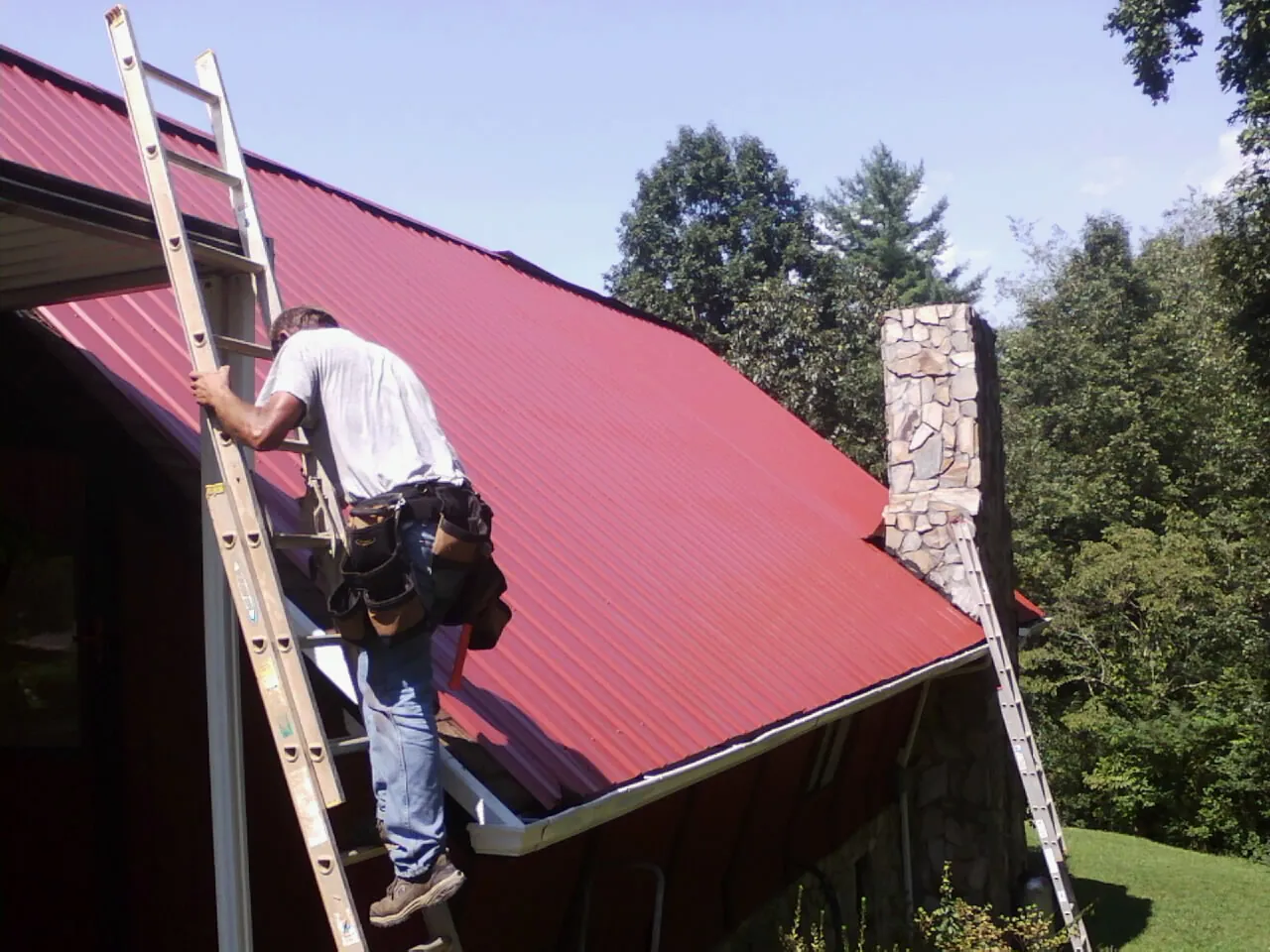 Roofer on a ladder beside a red metal roof and stone chimney — Soffit & Fascia Repair by Roofing Brothers in Webster Springs